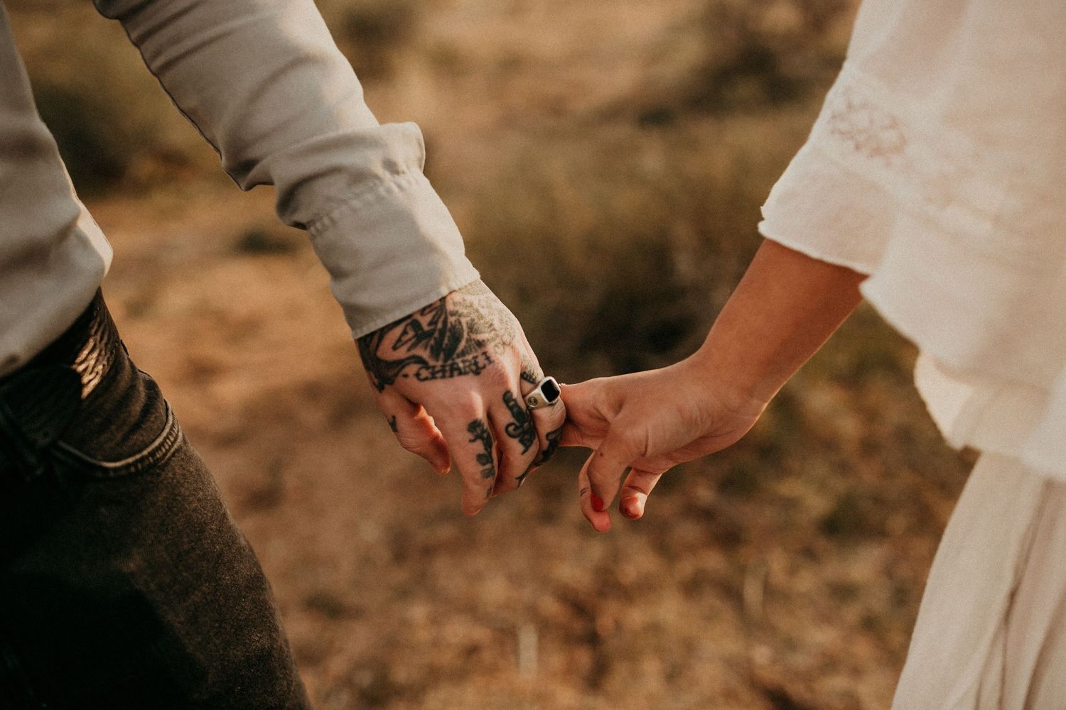 Two hands with tattoos touching intimately against a desert backdrop.