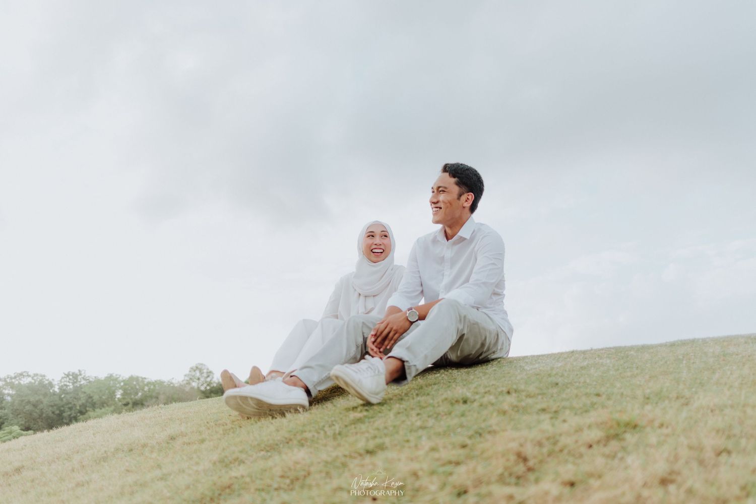 A couple in white clothing sits together on a grassy hilltop against an overcast sky.
