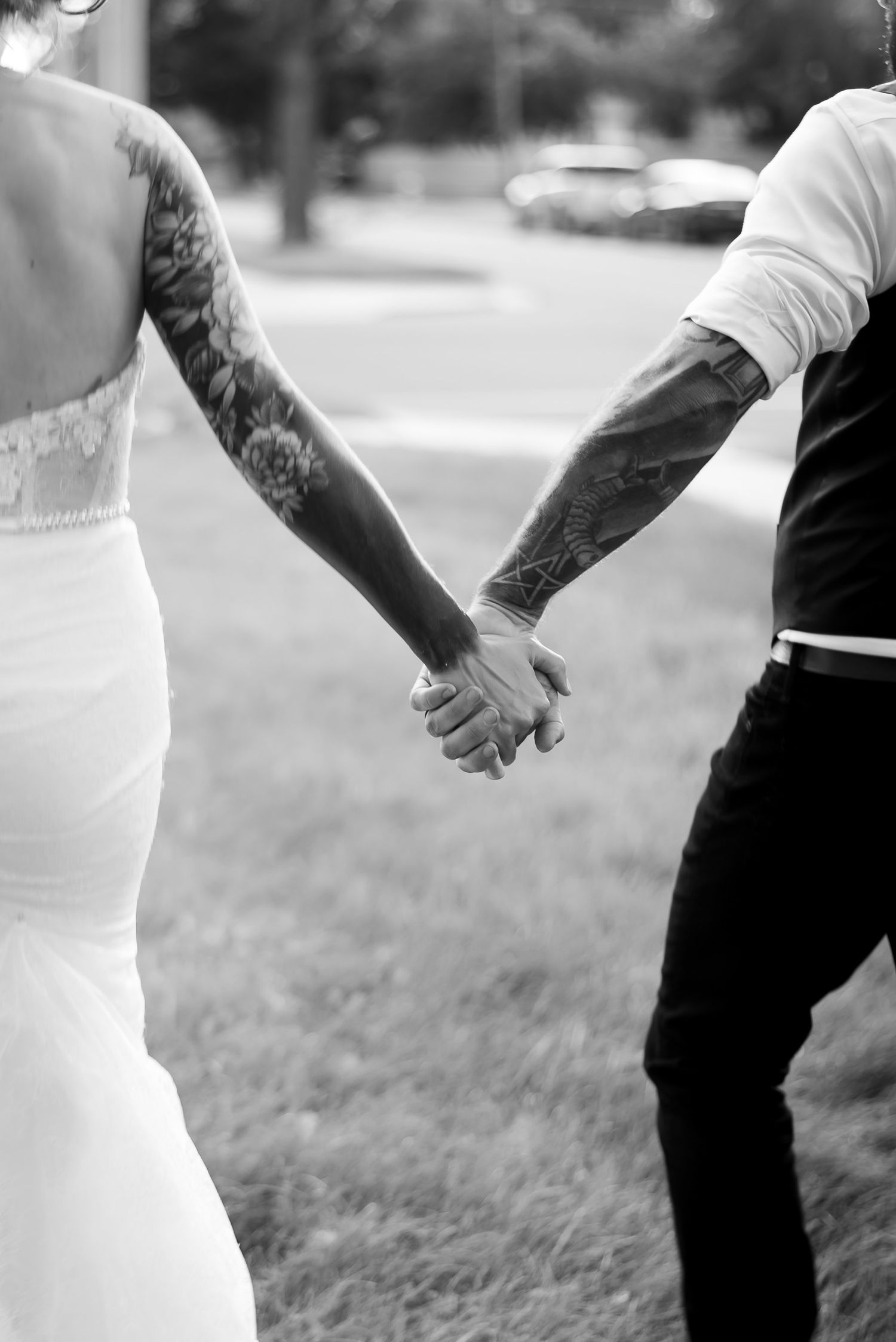 Black and white photo of tattooed couple holding hands in wedding attire outdoors.