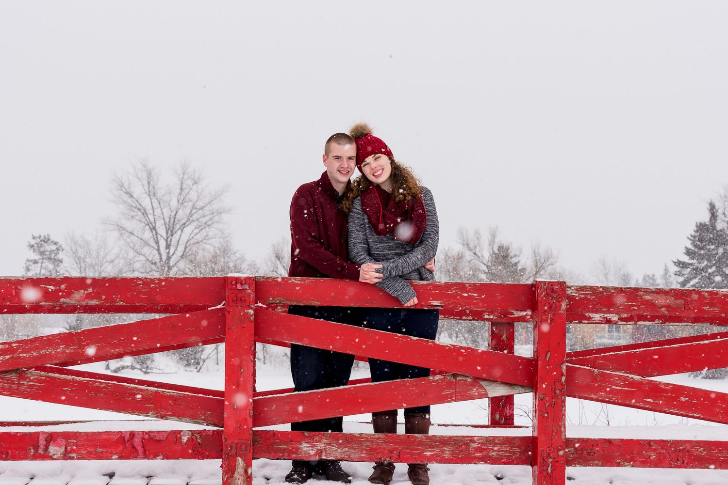 Couple standing on red wooden fence in snowy winter landscape.