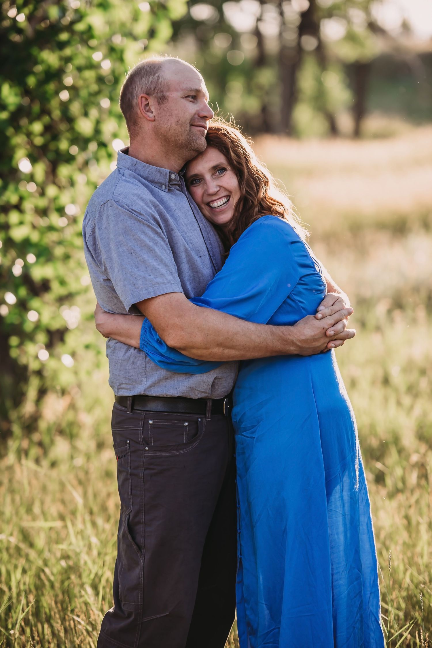 A couple embraces lovingly outdoors in a natural setting with golden sunlight filtering through trees in the background.