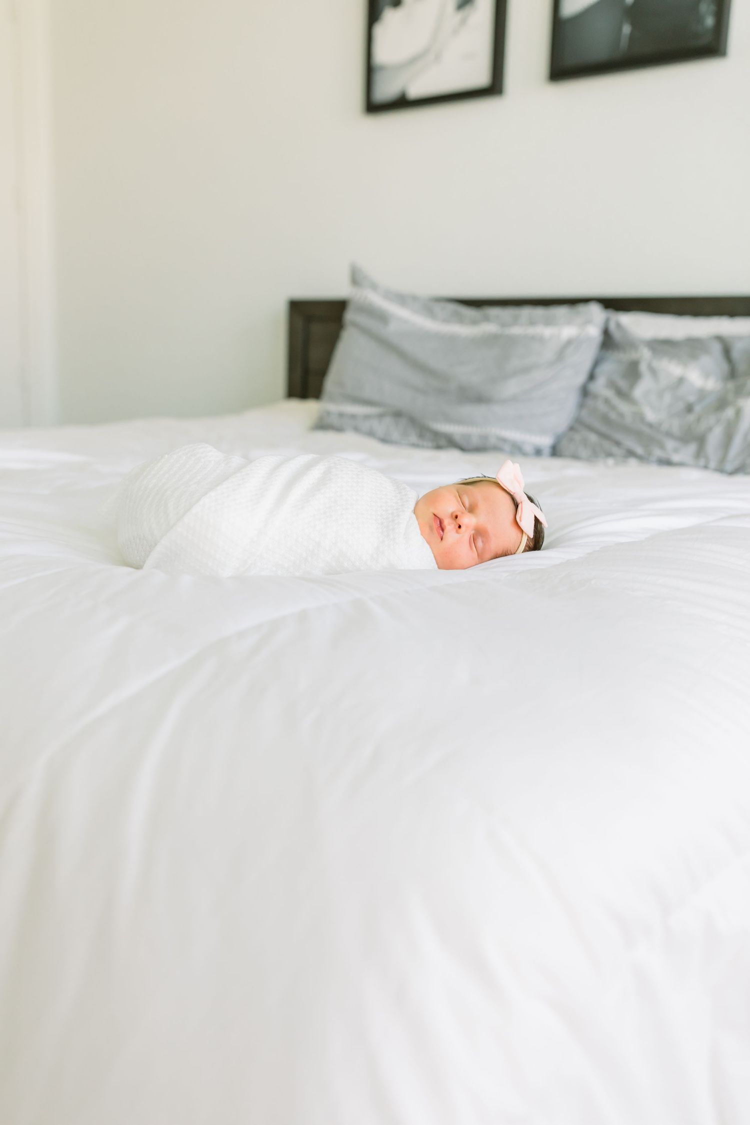 A sleeping newborn baby wrapped in white on a bed with grey pillows.