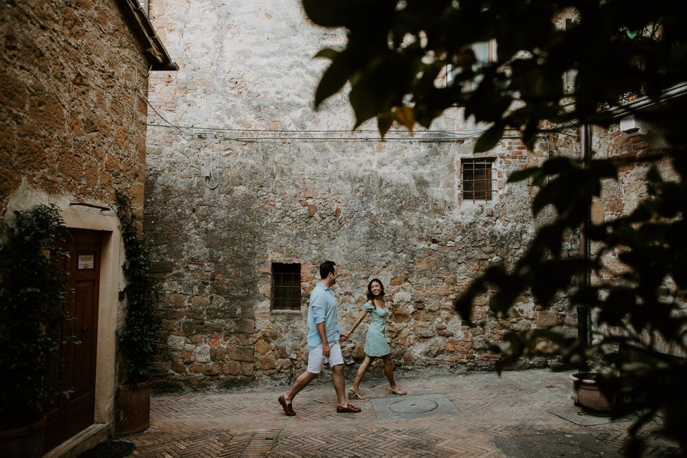 A couple walking together in a rustic stone courtyard with natural lighting and greenery.