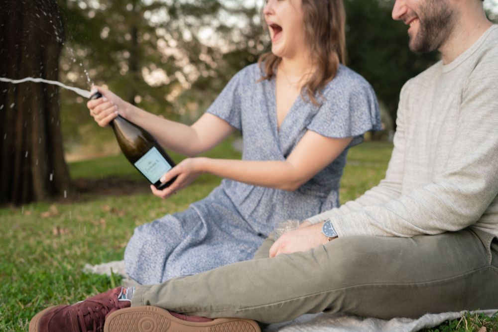 A couple celebrates by popping open a bottle of champagne during an outdoor picnic.