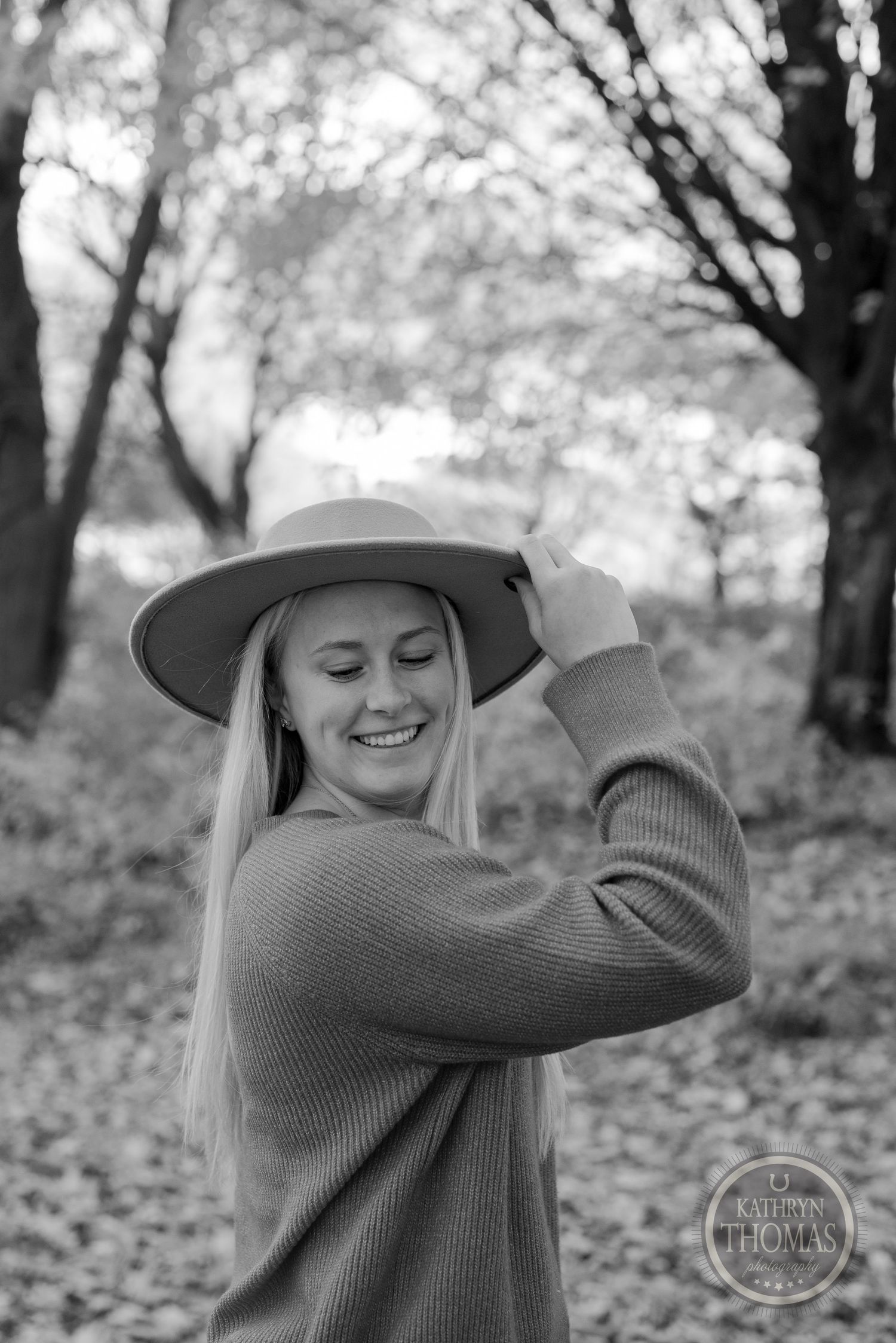 Black and white portrait of someone in a sweater and hat smiling among autumn trees.