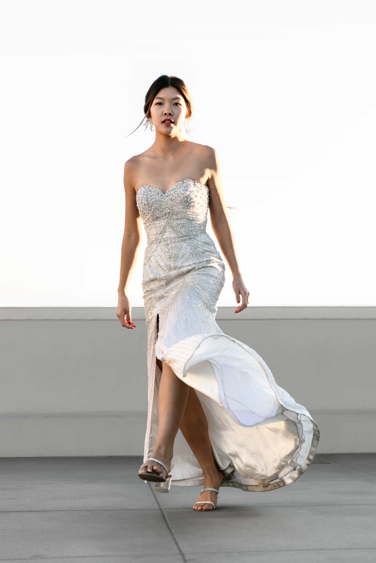Model walks in strapless crystal-embellished mermaid gown with flowing slit skirt against bright sky.