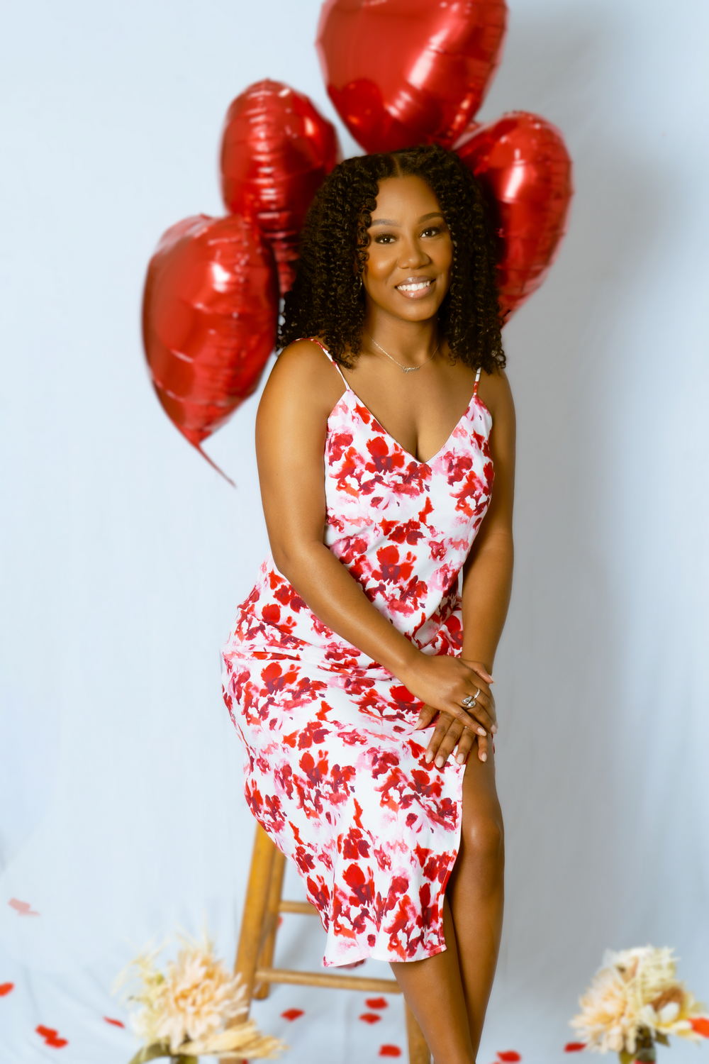 A person in a red and white floral dress poses with heart-shaped balloons against a white background.