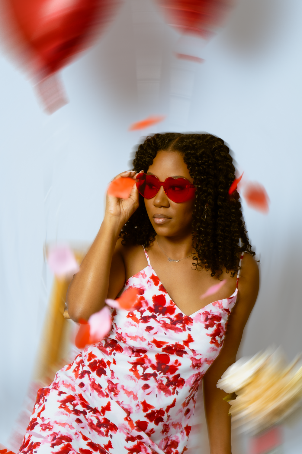 Someone wearing red sunglasses and a floral dress poses with heart balloons in a dreamy setting.