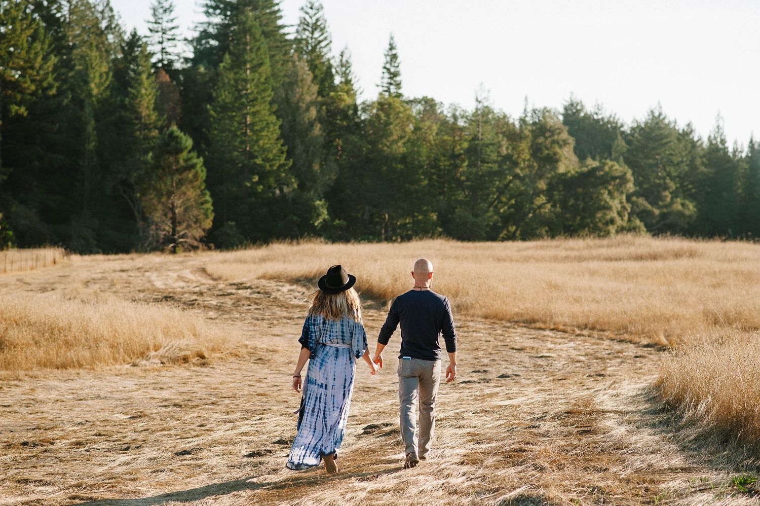 Couple walking hand in hand through golden field with forest backdrop.
