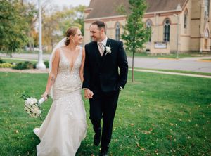 A couple in wedding attire walks together on grass with a church in the background.