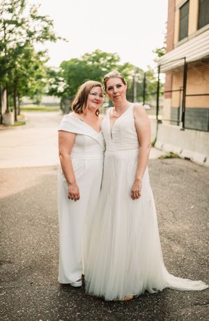 Two brides in elegant white dresses pose together on an urban street.