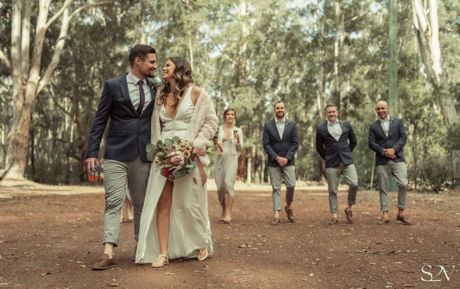 Wedding party walks through eucalyptus forest with groomsmen following in matching attire at sunset.