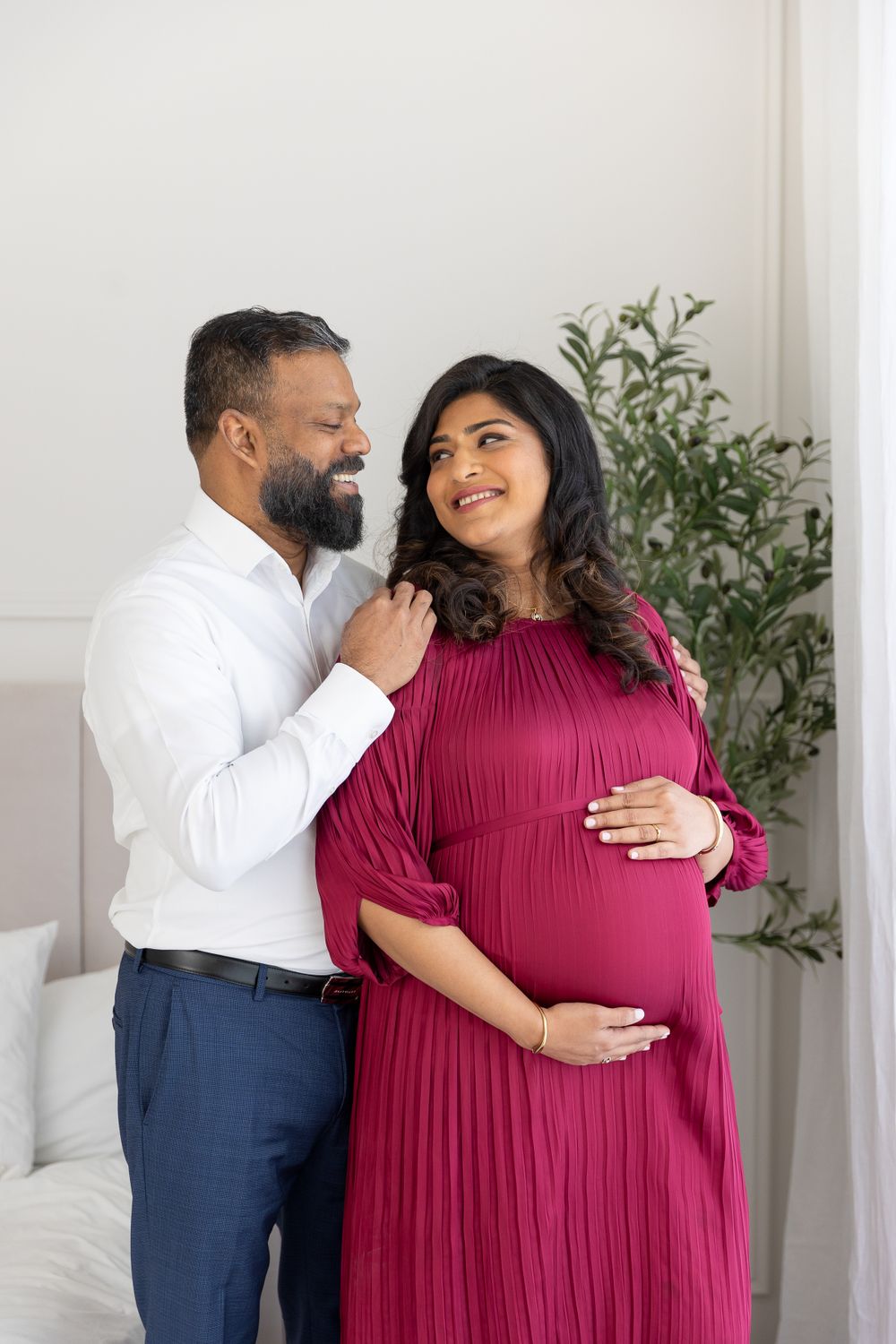Happy pregnant couple embracing by the window in a neutral studio space.