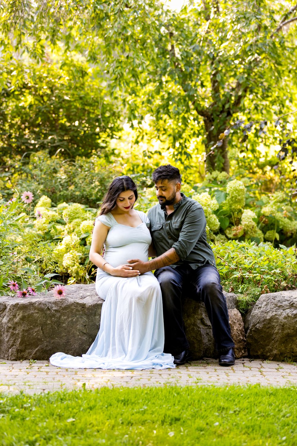 A couple sits on rocks in a lush garden with flowers and greenery during a maternity photo session.