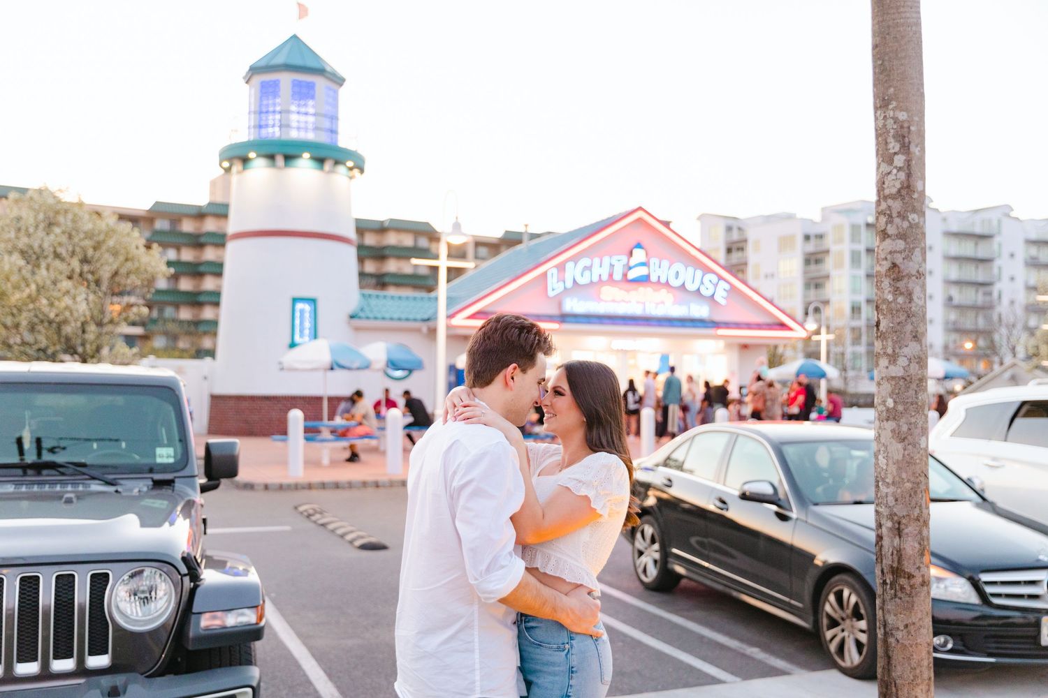 Engaged couple sharing a kiss in front of colorful Lighthouse restaurant sign at golden hour.