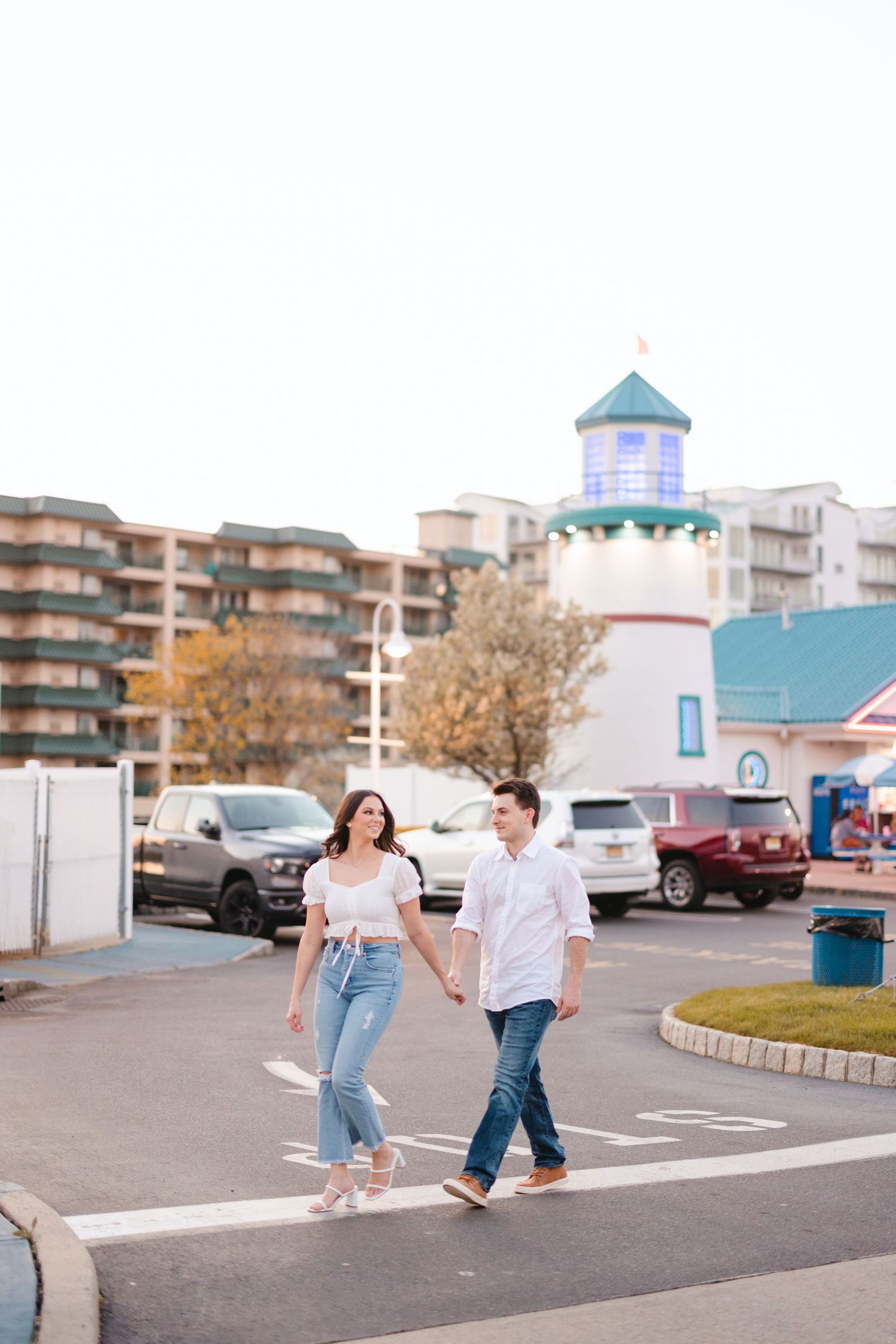 Engaged couple in white tops and jeans holding hands crossing parking lot near lighthouse landmark.