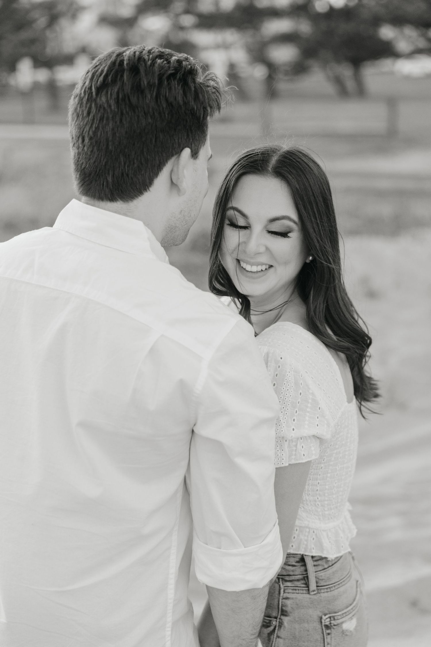 Black and white photo of a smiling brunette woman in white top gazing at her partner during engagement shoot.