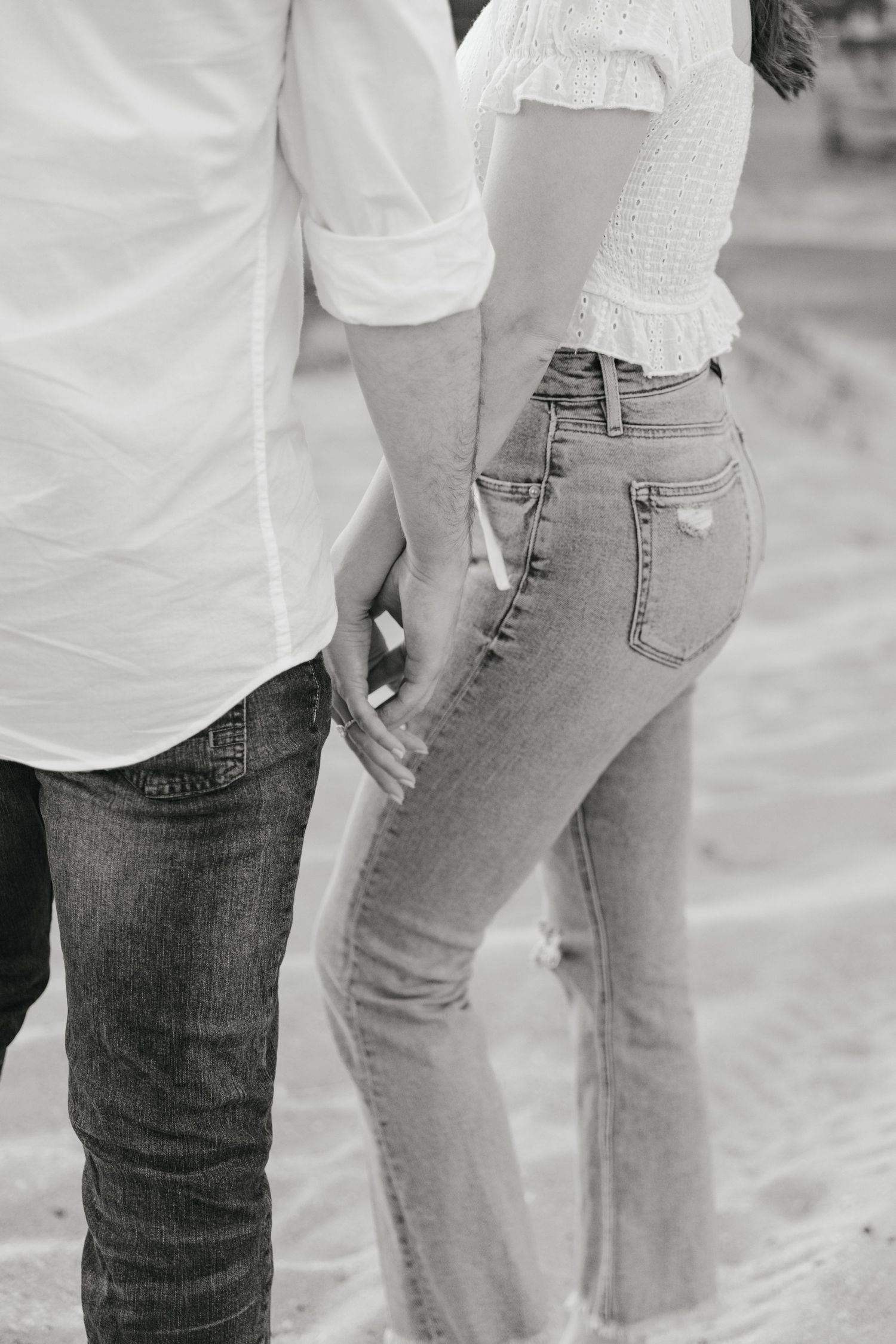 Black and white photo of couple holding hands on beach, both wearing white shirts and jeans.