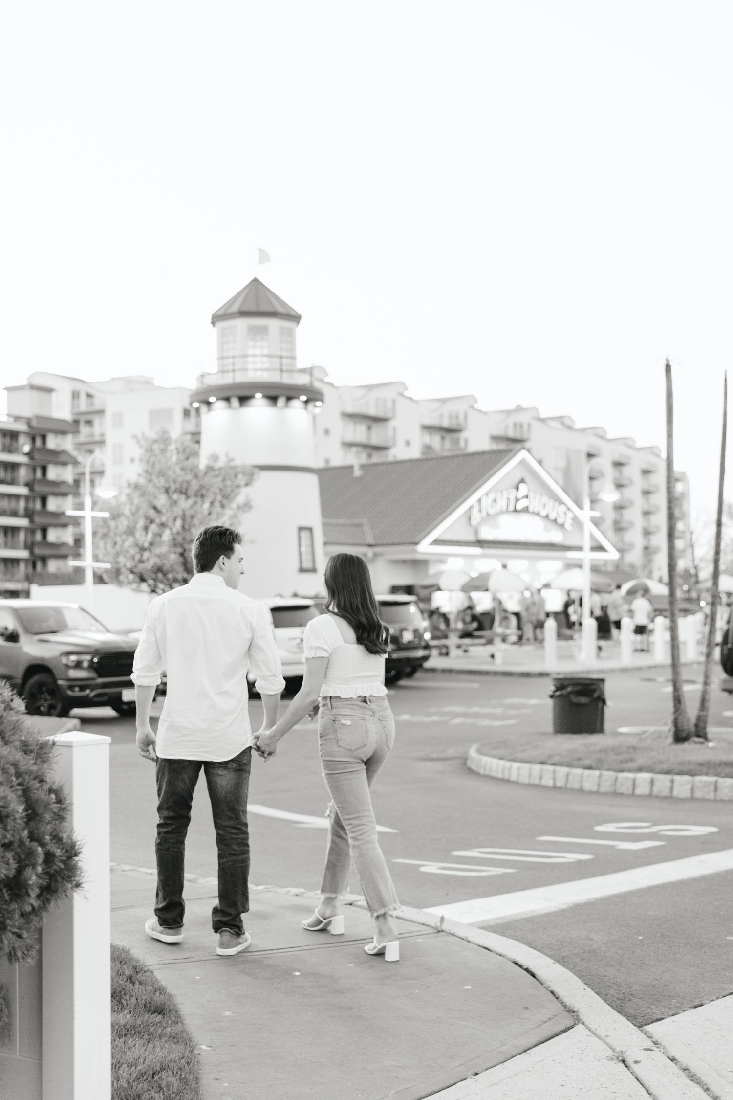 Black and white photo of couple holding hands walking toward Lighthouse restaurant during engagement session.