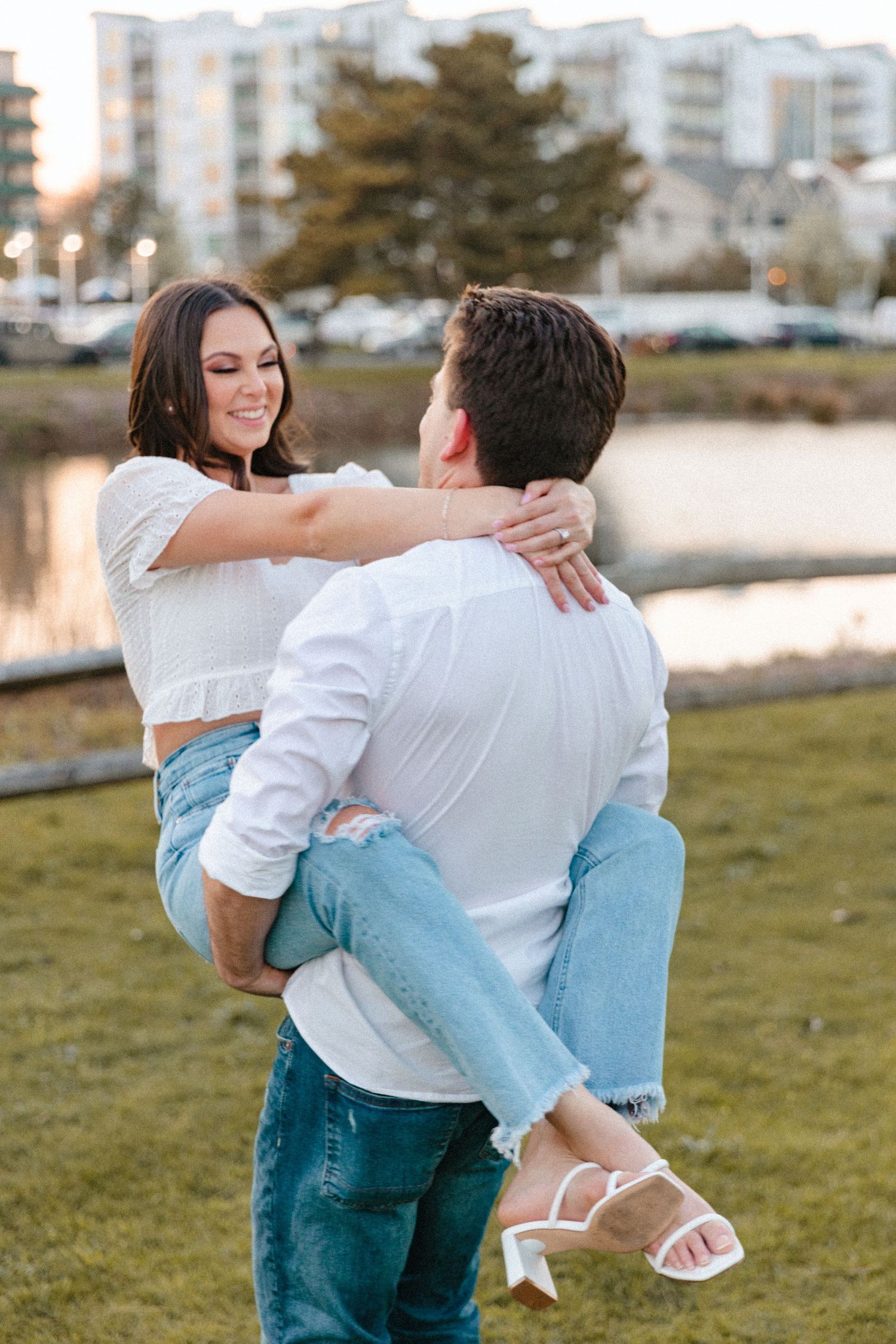 Man lifting woman in white crop top and jeans during couples engagement photo session near waterfront.