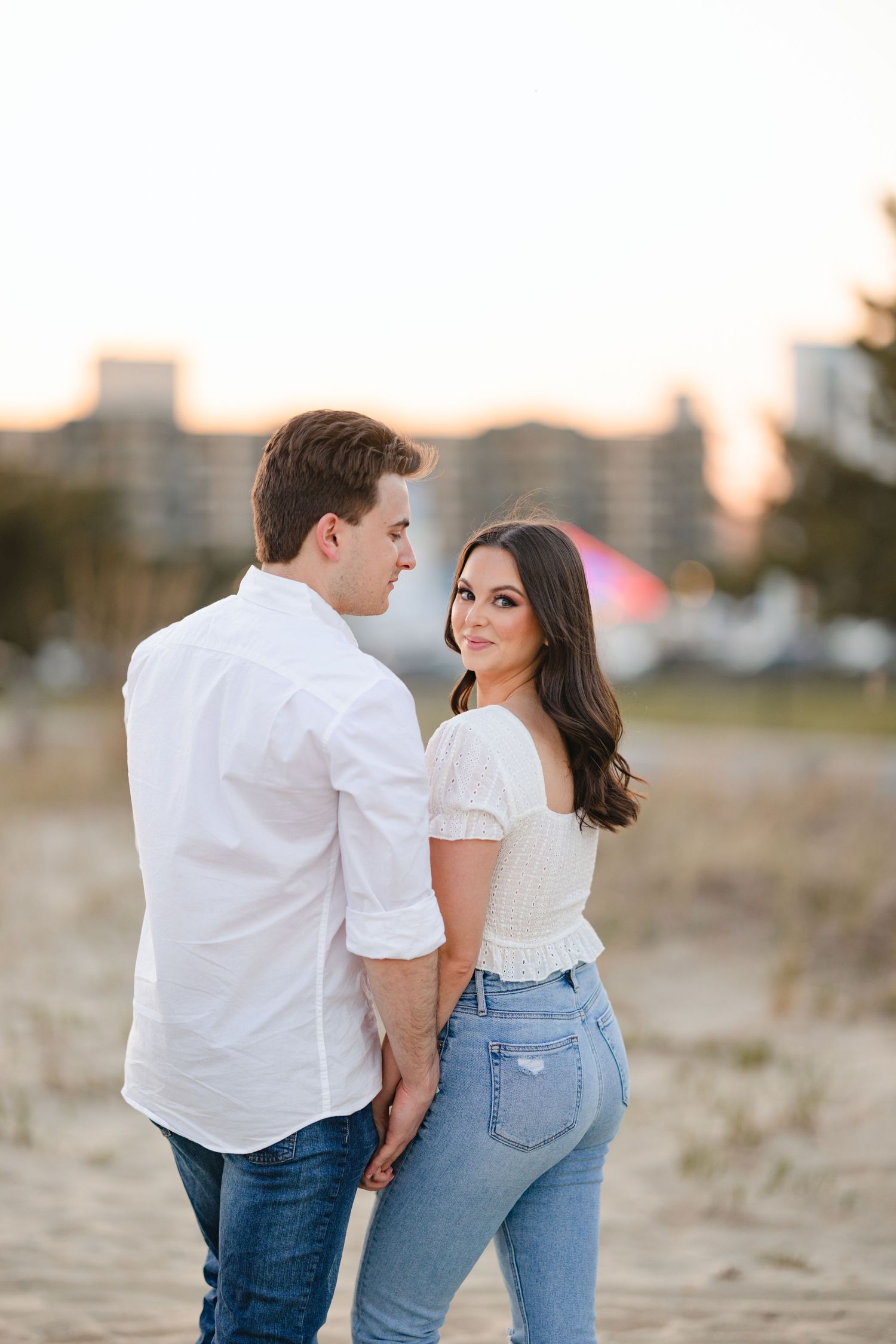 Engaged couple holding hands on sandy beach at sunset, woman in white top and jeans looking back.