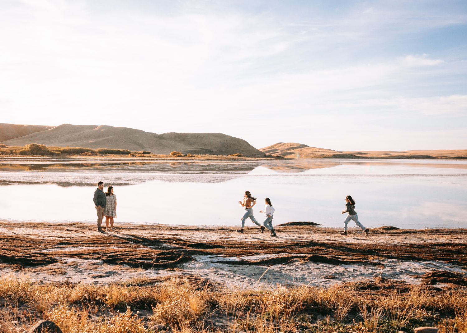 Active family running along a scenic lakeside with rolling hills reflecting in calm water.