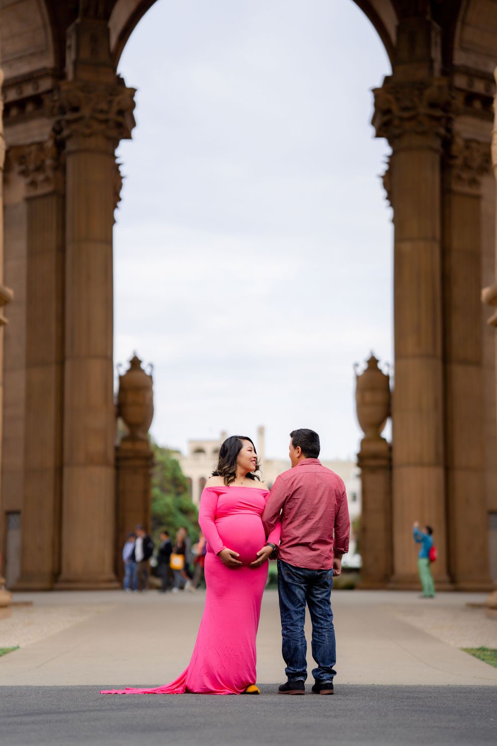 A couple stands beneath an ornate stone archway at Palace of Fine Arts with the woman wearing a flowing pink dress.