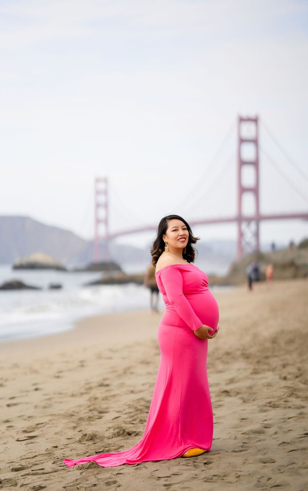 A maternity portrait on a beach showing a woman in a flowing pink dress near the Golden Gate Bridge.