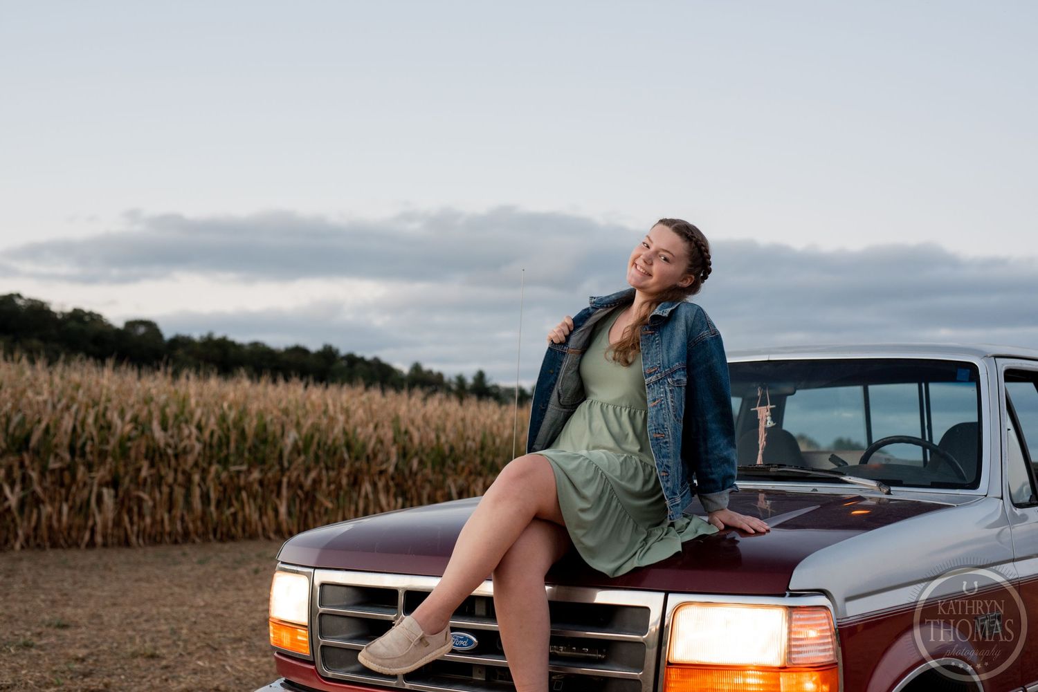 Casual portrait sitting on vintage truck hood at dusk with cornfield in background.