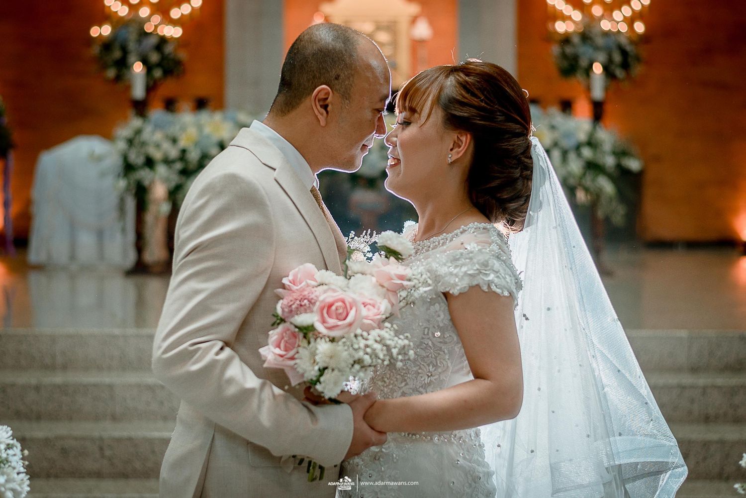 Romantic moment between newlyweds with pink bouquet and ornate floral backdrop.