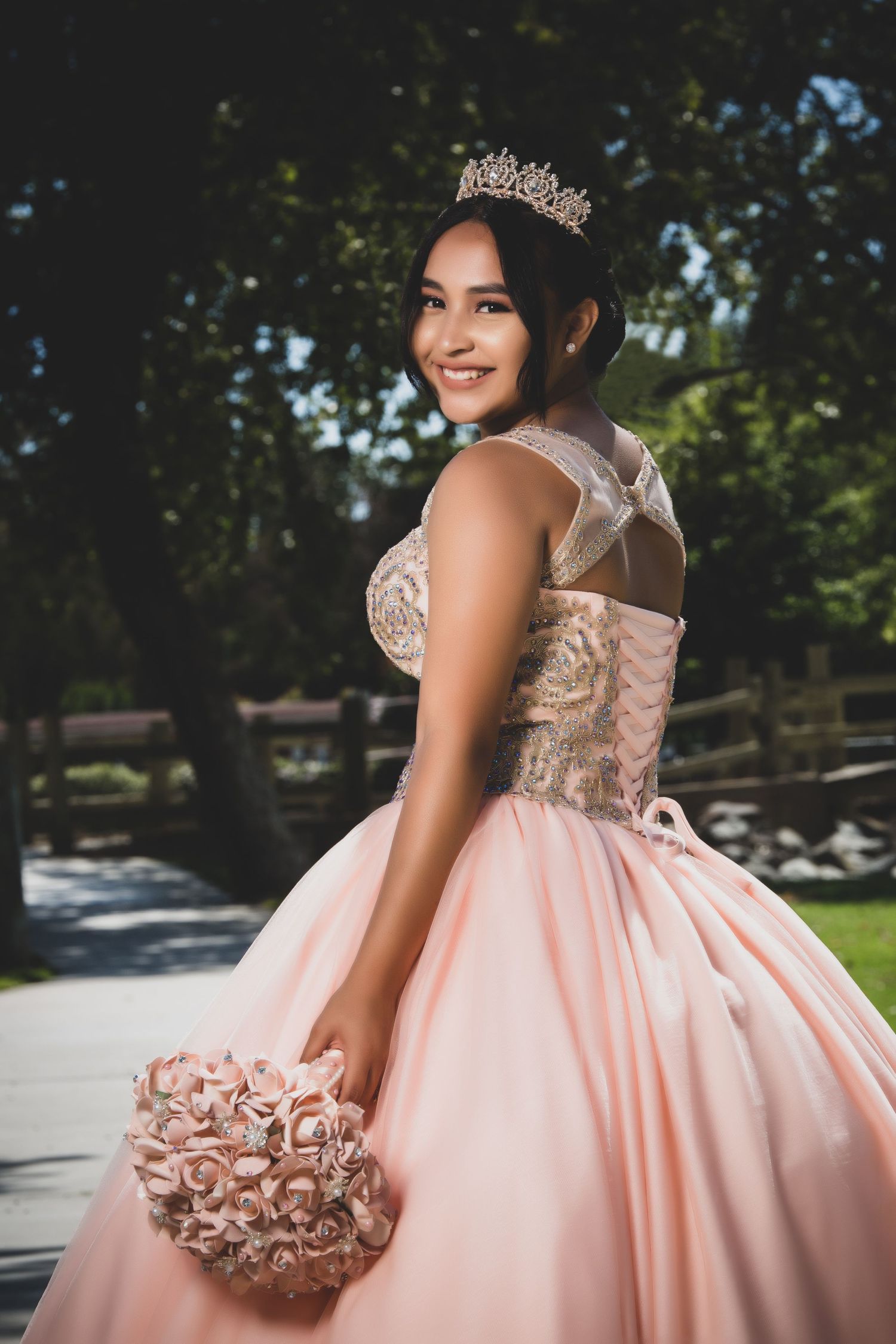 A person in a pink quinceañera ballgown with a sparkly crown poses outdoors holding a rose-colored bouquet.