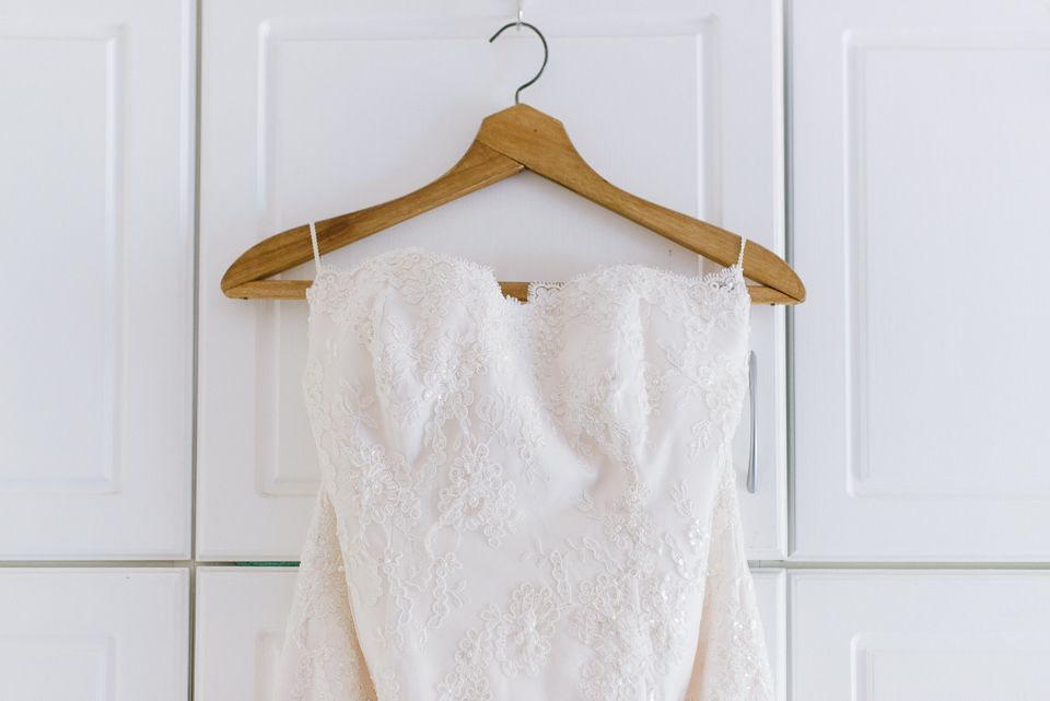 White lace wedding dress hanging on wooden hanger against white paneled wall.
