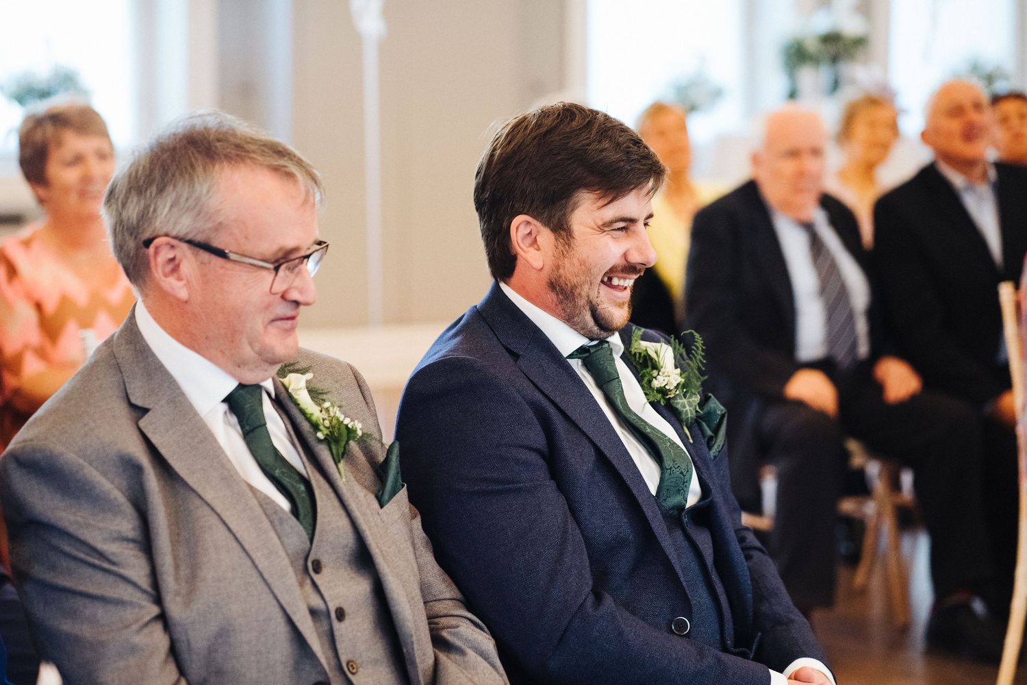 Two guests watching a wedding ceremony in formal attire with boutonnières in a bright indoor venue.