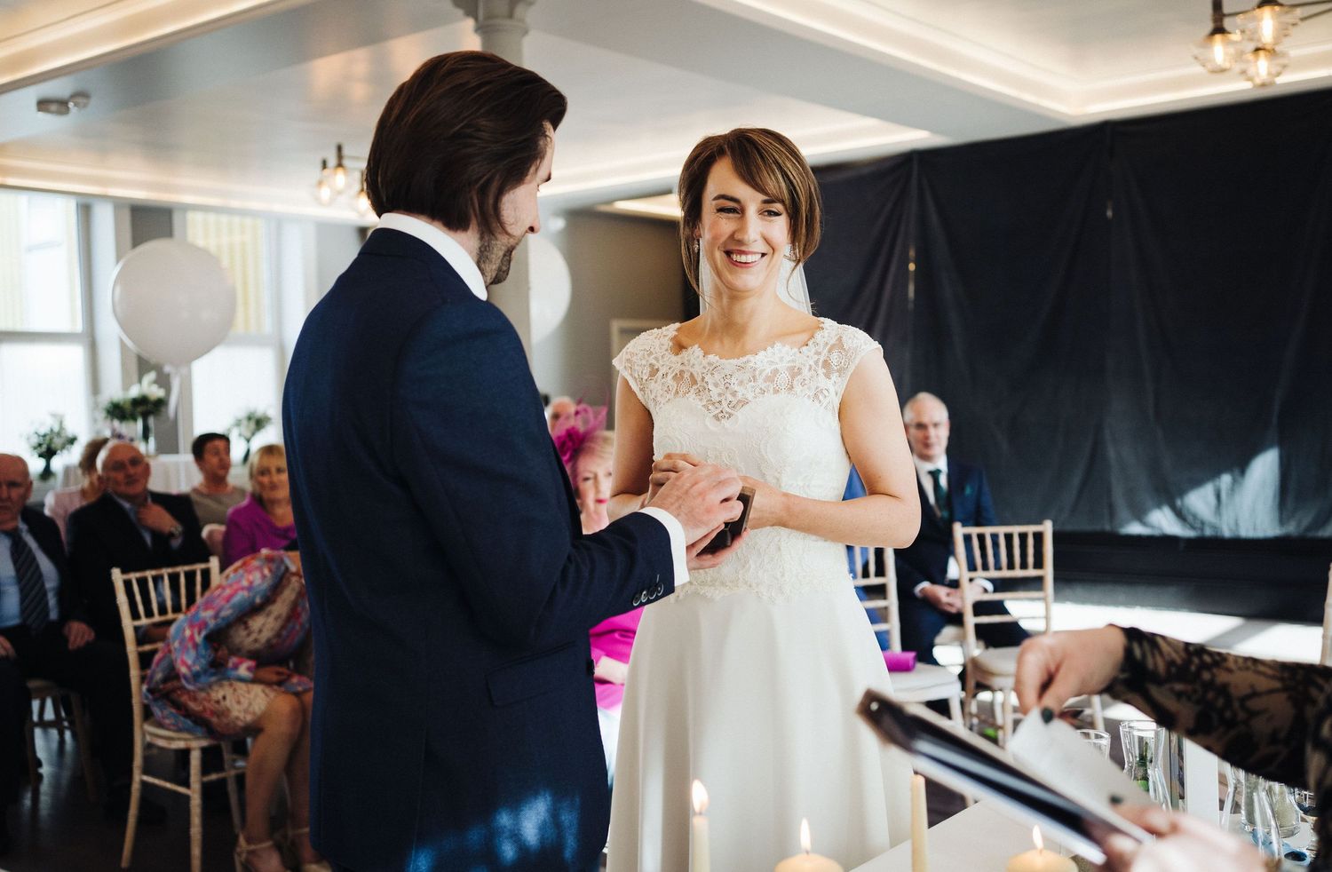 A couple exchanges rings during their wedding ceremony in an elegant indoor venue with decorative lighting.