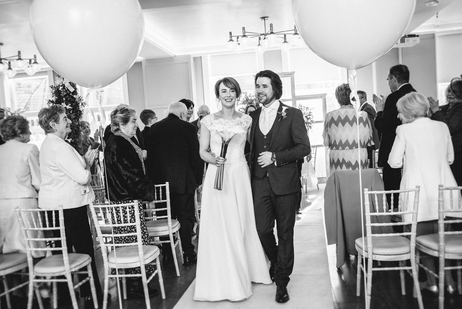 Black and white photo of a couple walking down the aisle with large white balloons and chiavari chairs.