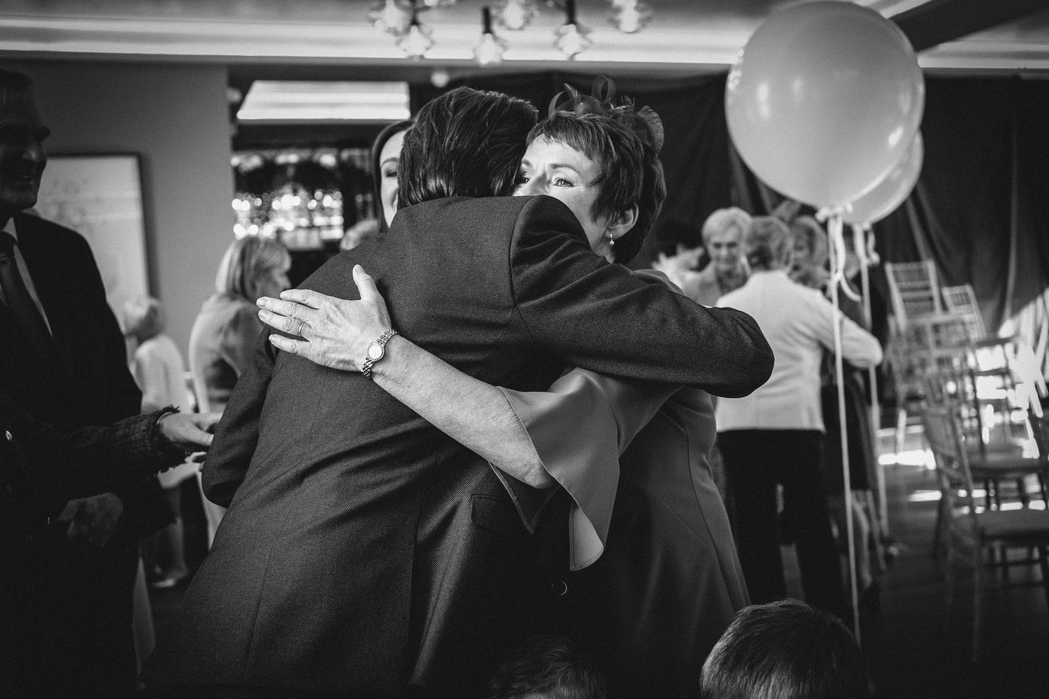 Black and white photo of an emotional embrace during a wedding reception with balloons in the background.