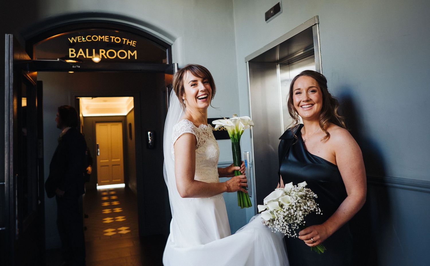 Outside a ballroom entrance, guests pose with bouquets and formal attire.