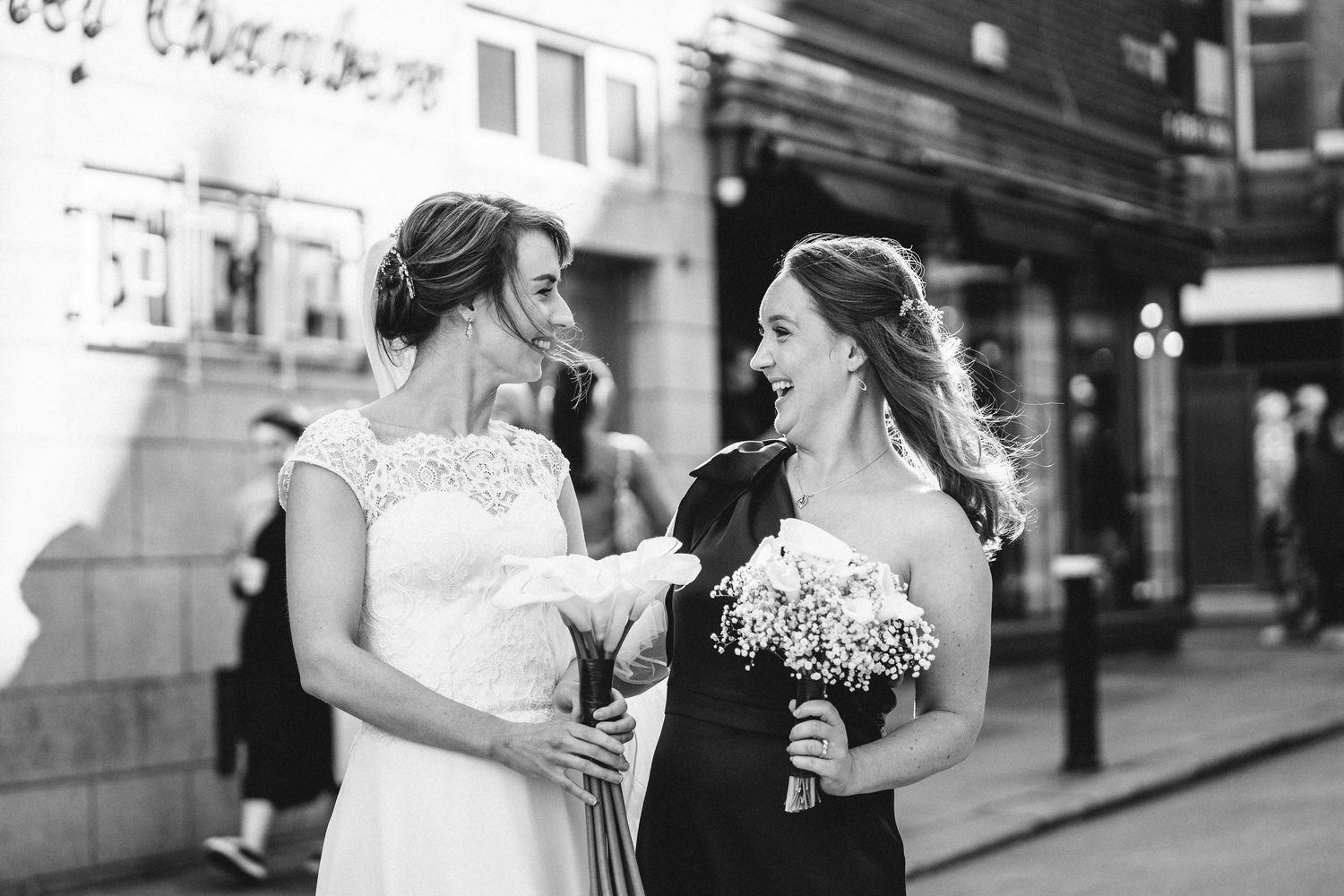 Two figures in formal attire share a candid moment together on a city street, one holding a bouquet.