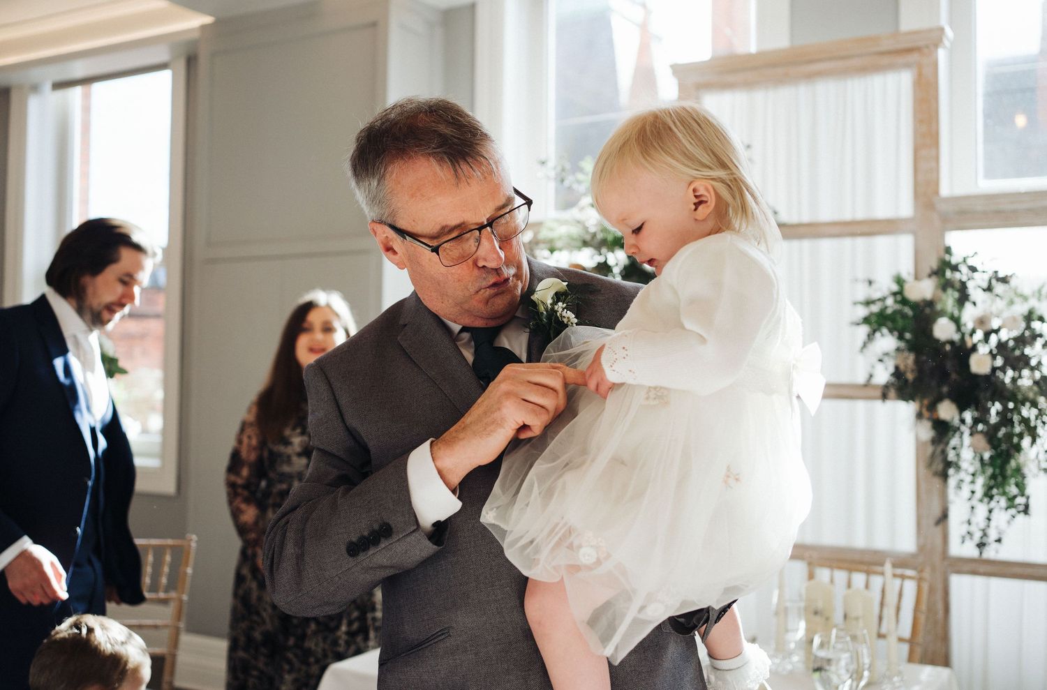 A tender moment at a formal event as a man in a gray suit holds a small child in white.
