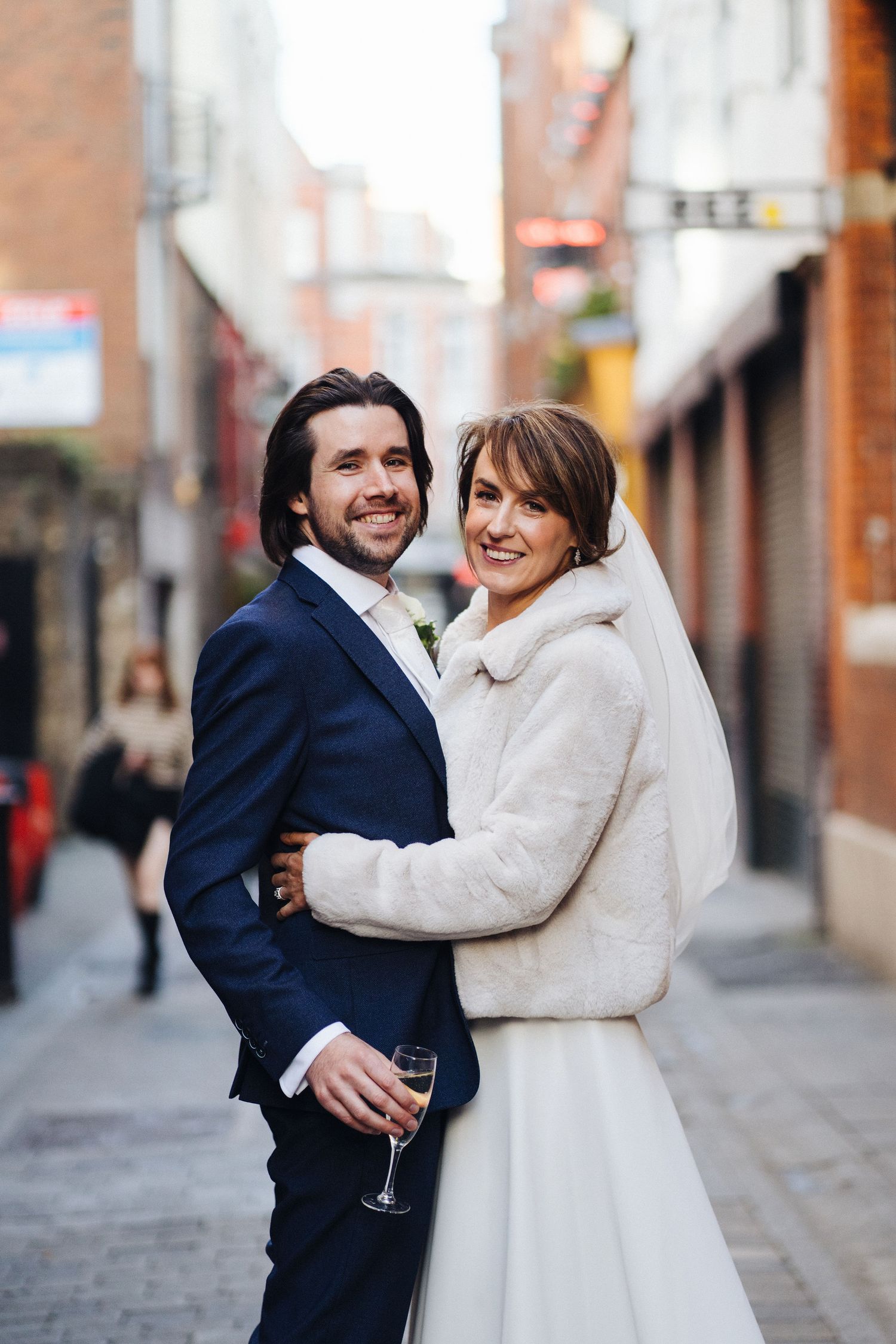 A couple embraces in an urban alleyway, with one holding a champagne glass.