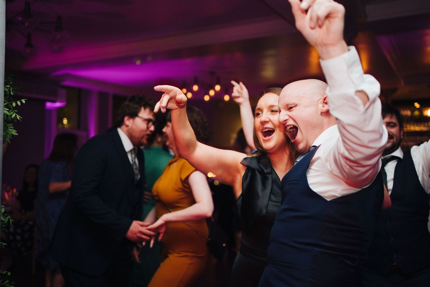 Party guests energetically dancing and celebrating at a reception with purple lighting.