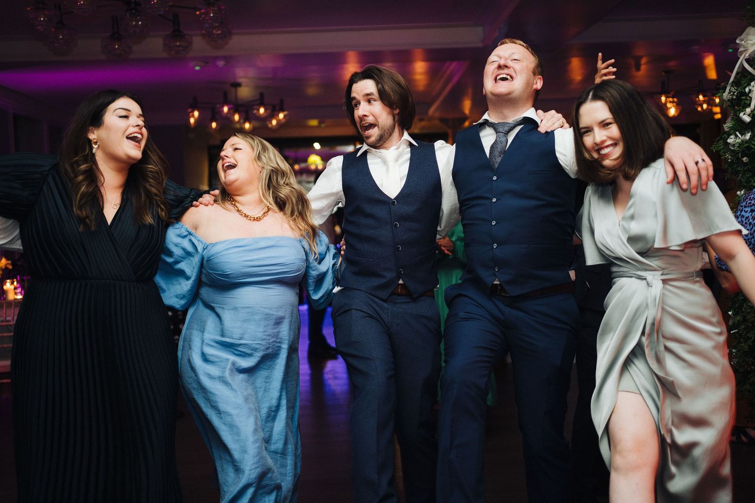 Group of wedding guests laughing together on dance floor with purple lighting.
