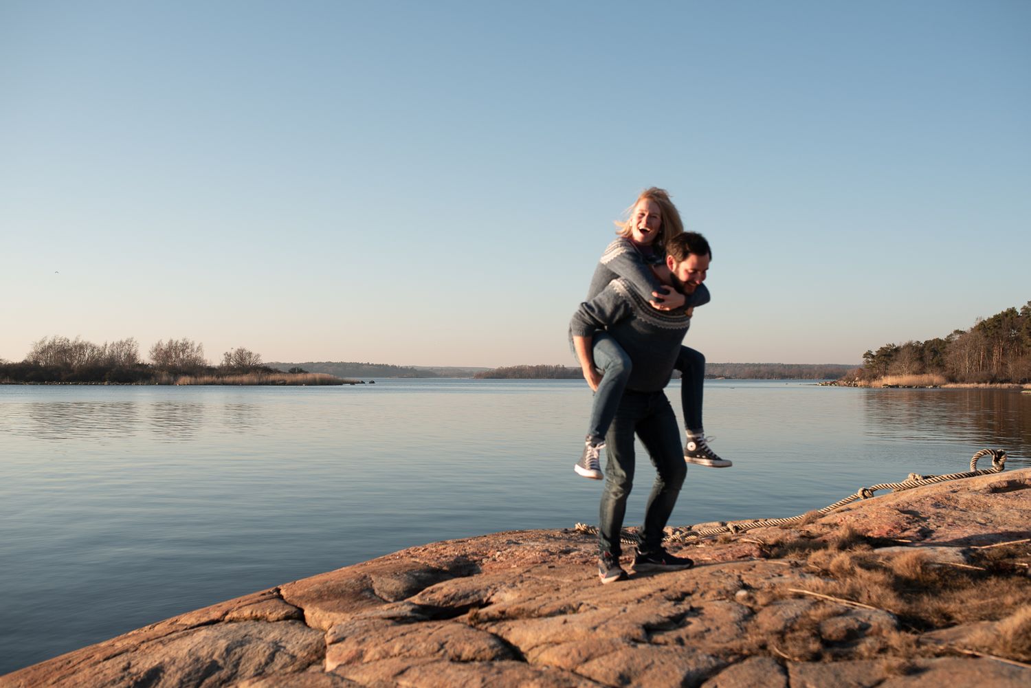 Romantic piggyback ride on rocky coastline with serene water view.
