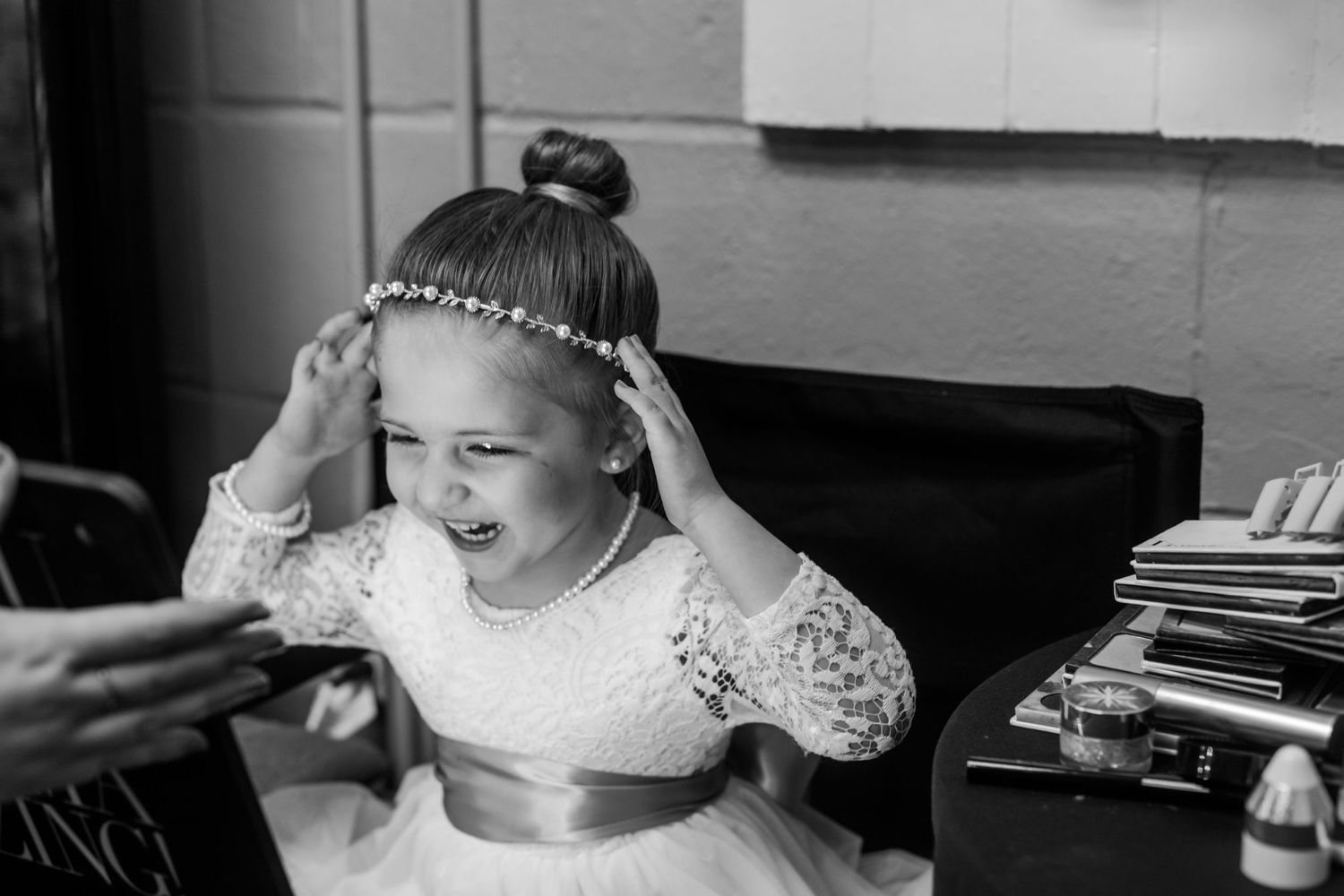 Black and white photo of a happy flower girl in white lace dress and pearl headband during wedding preparations.