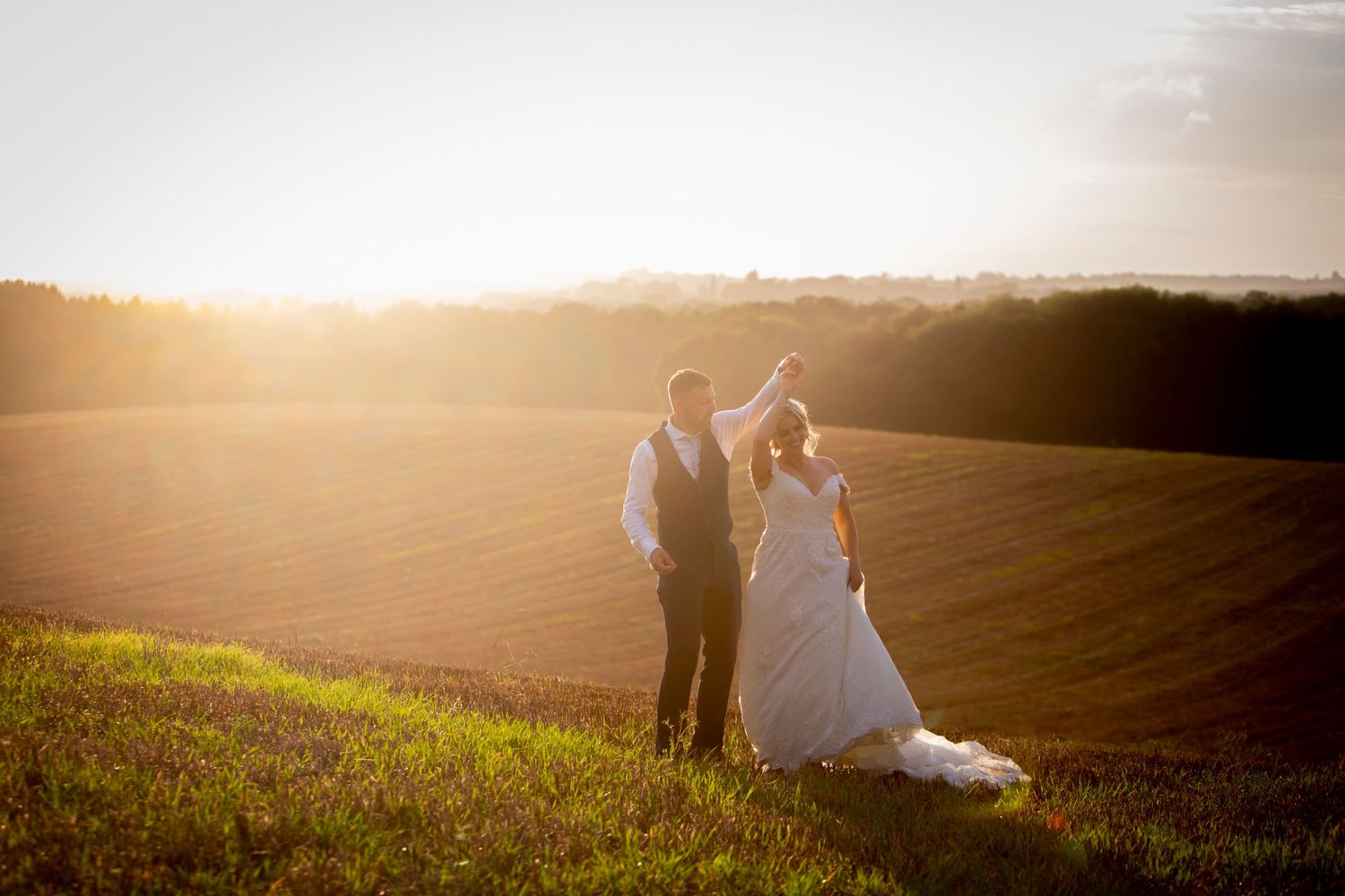 Bride and groom silhouetted against golden sunset over rolling fields.