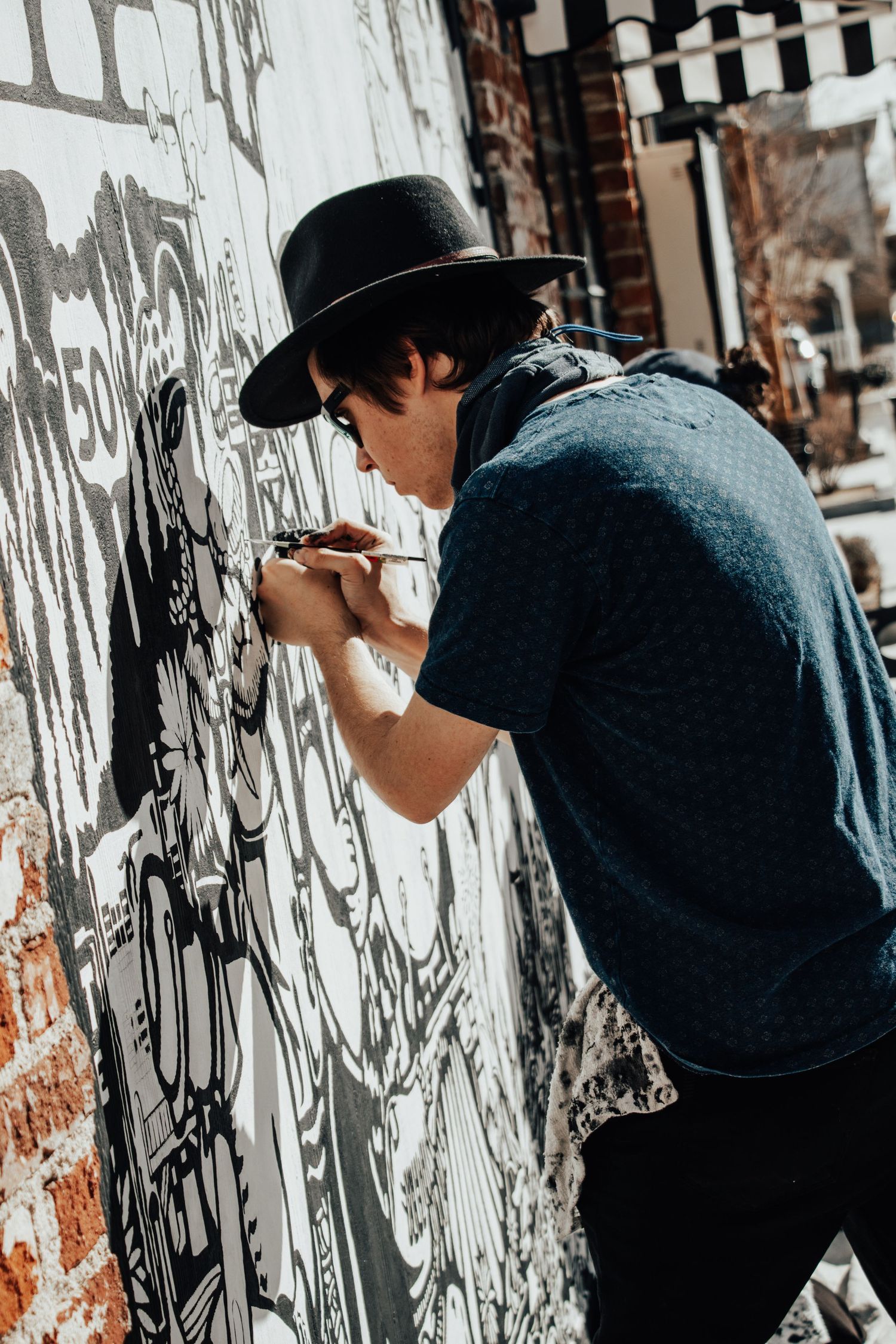 Artist in hat working on detailed street art mural on white urban wall.
