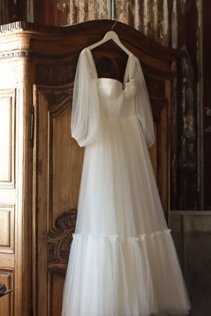 Cora Emilie Media An elegant white wedding dress with puffy sleeves hanging on display against an ornate wooden wardrobe.