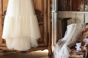 Cora Emilie Media Two flowing white wedding gowns displayed in a rustic room with vintage wooden furniture.