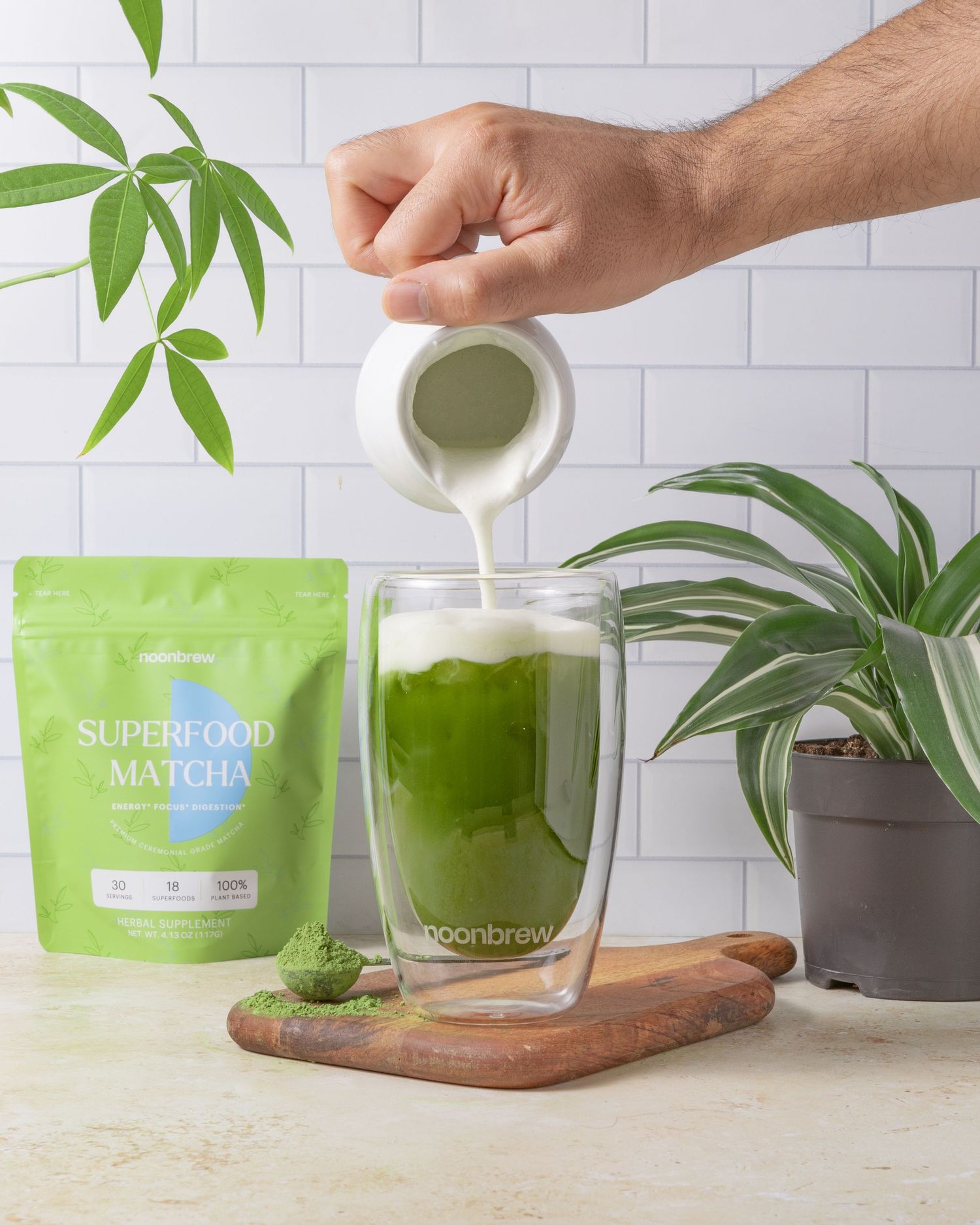 Hands pour matcha green tea powder from green package into a ceramic cup with plant decor on white counter.