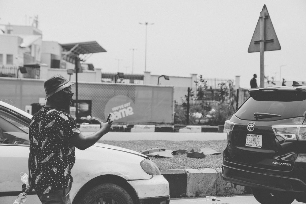 Black and white image of people gathered near parked cars in an outdoor setting.